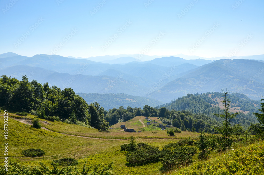 Obraz premium Mountain landscape with multiple layers of rolling hills and peaks fading into soft blue haze. Lush green trees and grassy slopes and valley with few small houses fill the foreground under clear sky