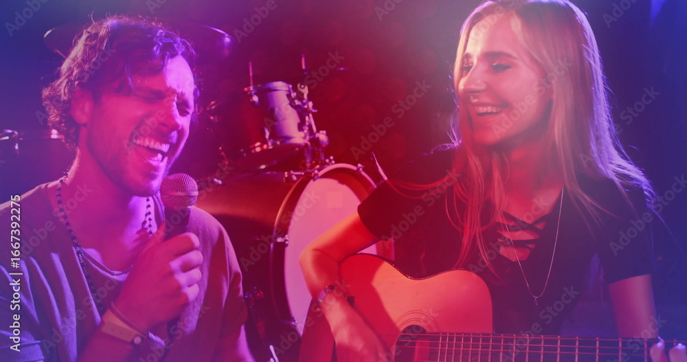 Naklejka premium Singing singer holding microphone as guitarist strumming guitar on stage, with drum kit and lights