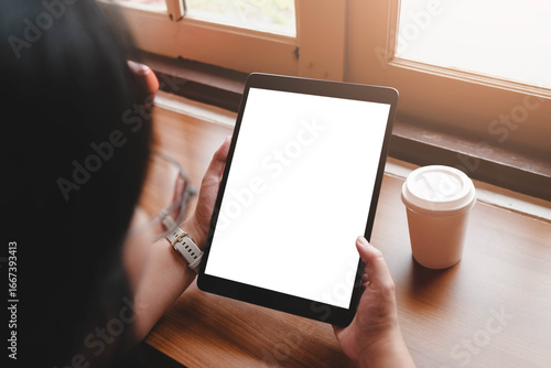 Over the shoulder view of a woman holding a tablet computer with a blank white screen, sitting by a window with a coffee cup. Perfect for mockups.