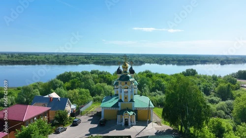 A historic yellow Russian Orthodox church with golden and green domes, located on a grassy hill with a white and green fence under a clear blue sky