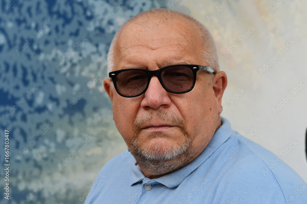 Obraz premium Portrait of an elderly gray-haired Caucasian man with a beard, wearing sunglasses and a blue T-shirt.