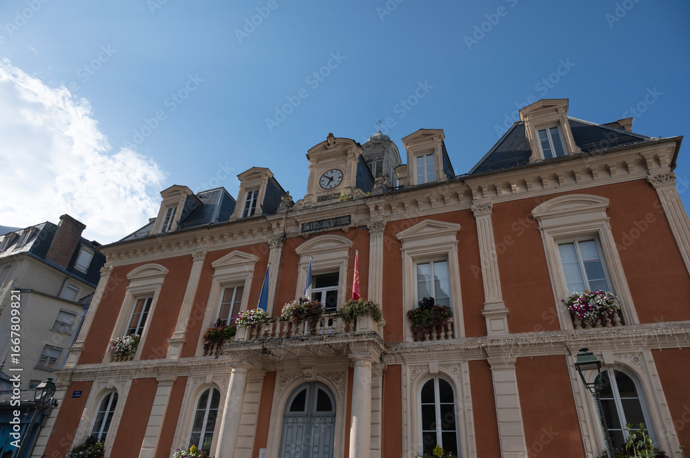 Fototapeta premium Cauteret's town hall building features elegant architecture with colorful flowers adorning the balconies, set against a backdrop of a brilliant blue sky and majestic mountains.