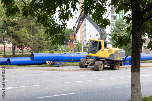 Wallpaper Mural Yellow excavator working near large blue plastic pipes at construction site in city street. Torontodigital.ca