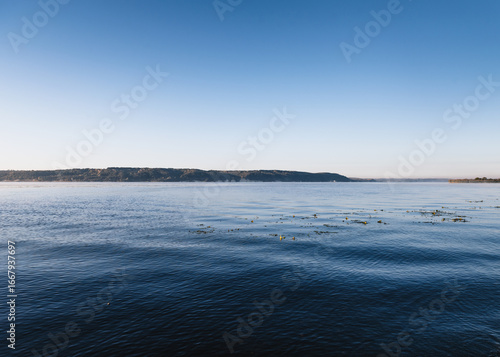 Lake in the morning. Camera close to calm water surface of the big lake shortly after sunrise. Gradient sky above montane bank of the river.