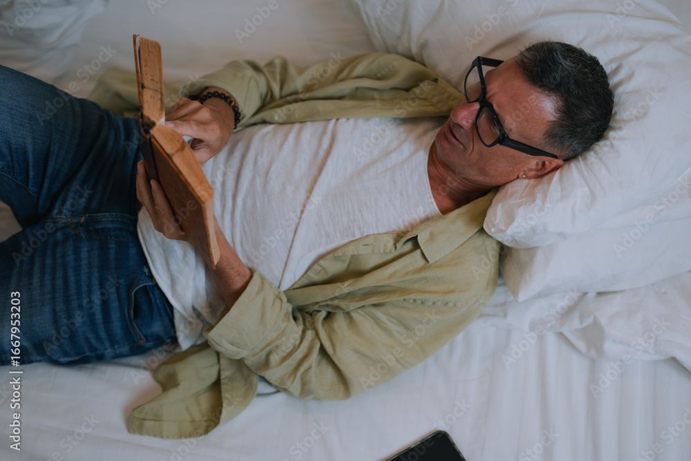 Fototapeta premium Mature man lying on a bed, wearing glasses and engrossed in a book, with a tablet resting on the sheets, enjoying a moment of relaxation and tranquility in his cozy bedroom