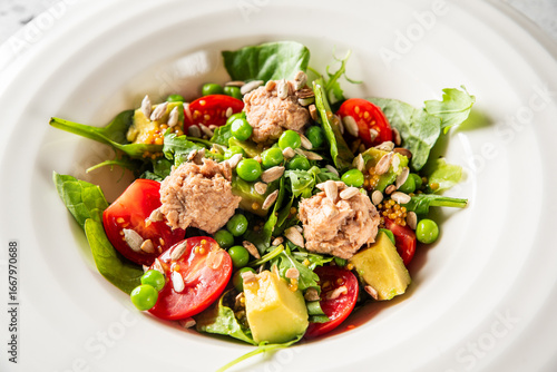 Healthy salad with cherry tomatoes, avocado, green peas, leafy greens, sunflower seeds, and tuna scoops in a white bowl. Fresh, colorful, and nutritious meal close-up.