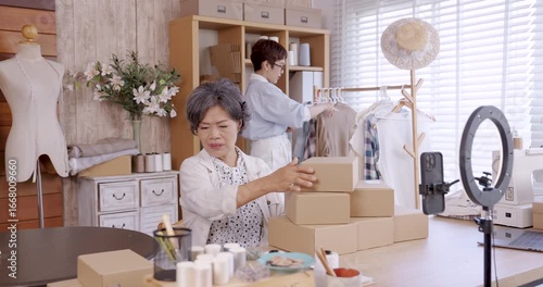 Senior asian woman business owner preparing packaging box while adult daughter checking clothes inventory in background, teamwork managing small fashion family business with e commerce shop
