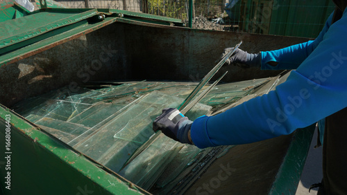 Hands in gloves disposing of old broken glass panes to waste containers in industrial recycling facility center. Processing recycled materials. Waste management, sustainability and safety 