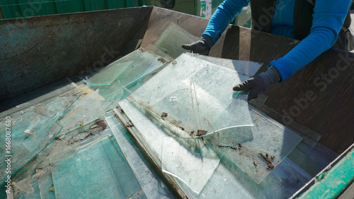 Hands in gloves disposing of old broken glass panes to waste containers in industrial recycling facility center. Processing recycled materials. Waste management, sustainability and safety 
