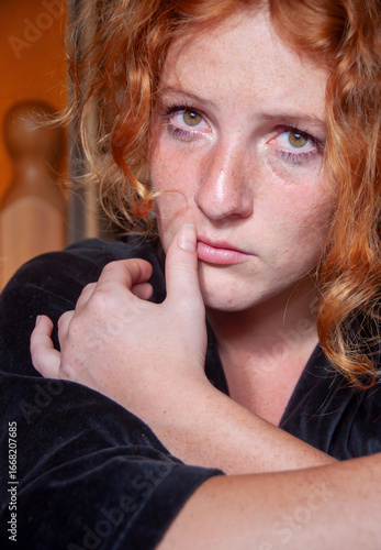 Close-up of a young red-haired woman with freckles and teary eyes, gazing vulnerably into the camera. Her hand touches her lips in a raw, emotional moment filled with tenderness and pain