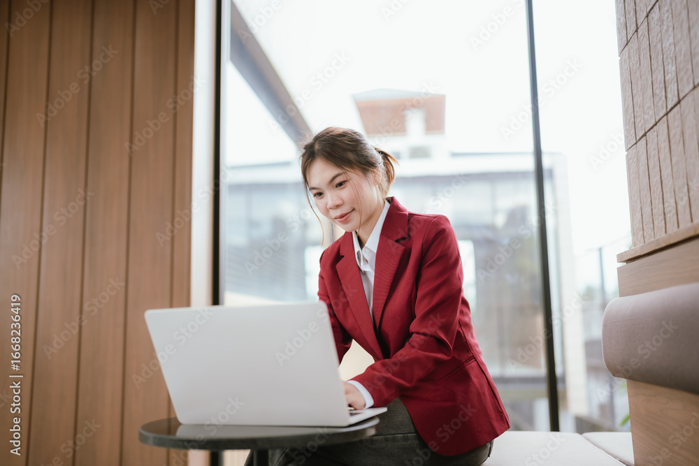 Fototapeta premium A smiling Asian businesswoman sits at her desk while working on her laptop. She is professional, cheerful and stylish. She efficiently manages digital projects, communications and online tasks.