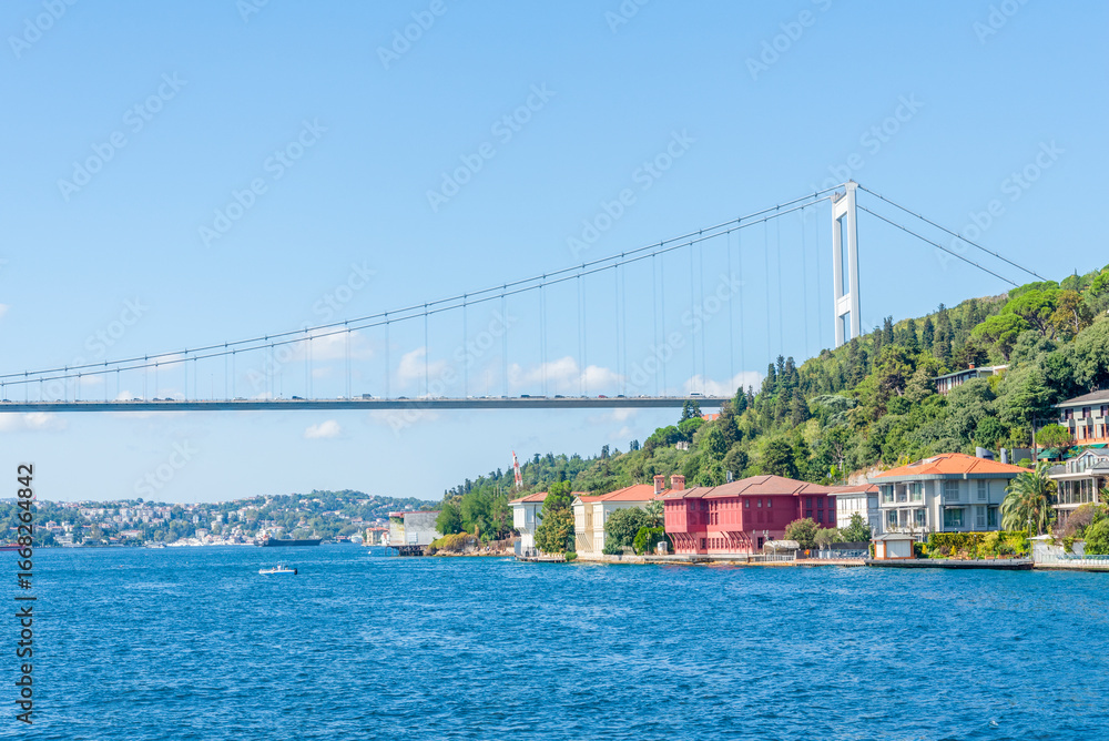 Fototapeta premium Bosphorus Bridge spans a serene waterway with charming waterfront homes in Istanbul on a sunny day