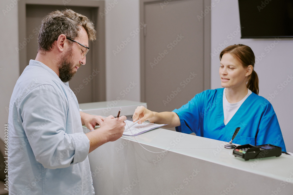 Obraz premium Caucasian middle aged man signing document at reception desk while Caucasian young adult woman in medical scrubs assisting him, both interacting in modern healthcare facility