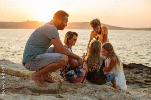 Family time outdoors in natural landscape. Parents with daughter and sons sitting on rocks by the seaside, enjoying a summer holiday at sunset.