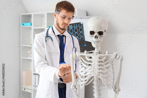 Young male doctor in white coat and smart glasses examining human skeleton mo...
