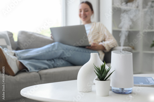 Photos Air humidifier with plant and vase on table against woman using laptop at home,