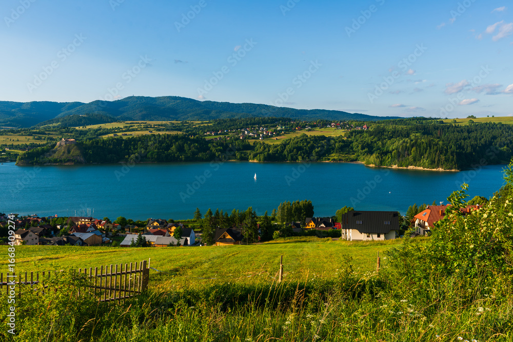 Fototapeta premium Charming village with scattered houses overlooking a calm blue lake, framed by distant hills and fields. A tranquil countryside scene in summer light.