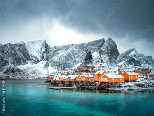 yellow huts of an Island with icy winter mountains in the background, foreground is clear blue water and there are dark storm clouds overhead. Winter in norway