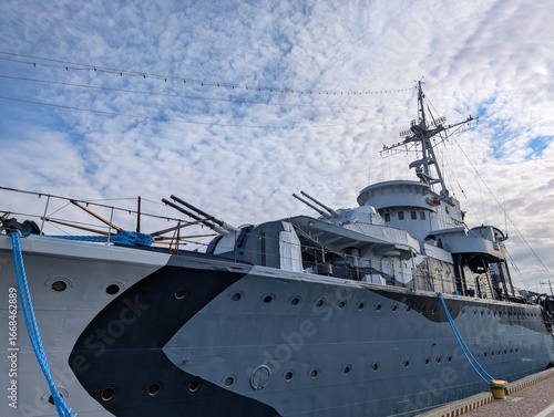 Historic naval battleship docked at the harbor on a cloudy day with visible artillery and modern equipment