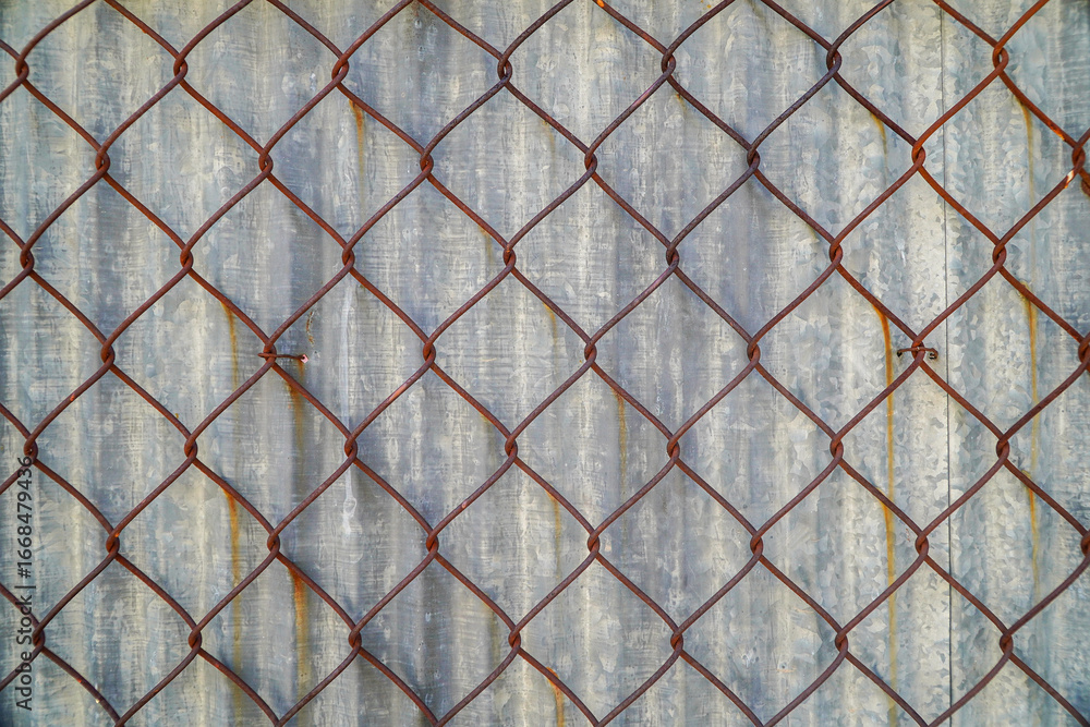 Fototapeta premium An aged chain-link fence with rust against a corrugated metal backdrop. The geometric pattern and weathered textures create an image suitable for industrial or abstract backgrounds.