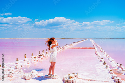 beautiful nature pink lake salt flats where salt is mined with soft pink water blue sky fluffy clouds and snow-white salt on the shore with a red-haired girl in a white dress looking into the distance
