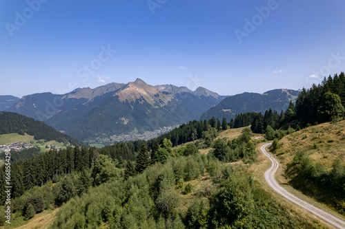 Aerial mountainous landscape with meandering path through lush green French Alps valley tourist destination winter sports near Les Gets village with rocky mountain top against a clear blue sky. 