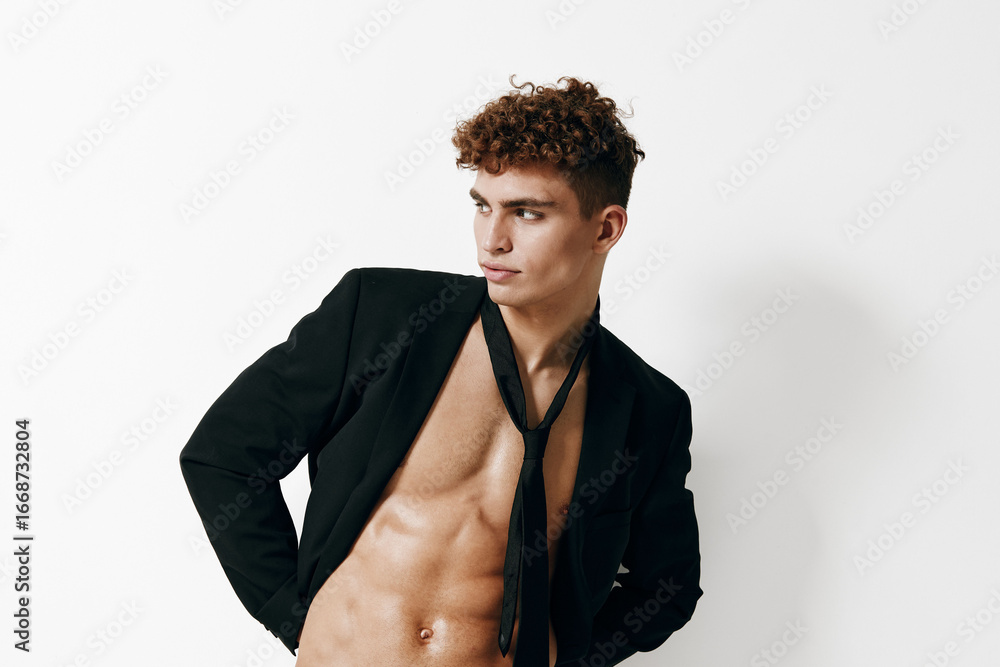 Obraz premium Stylish young man with curly hair posing confidently in a black blazer without a shirt, showcasing a modern and edgy look against a plain white background.