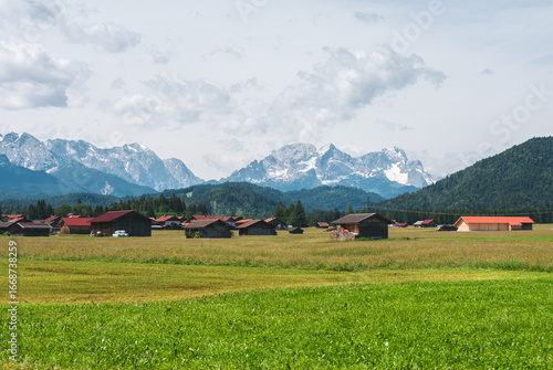 Traditional Bavarian barns and red-roofed farm buildings in green fields, with the snowy Alps rising in the background. A peaceful rural landscape in southern Germany.