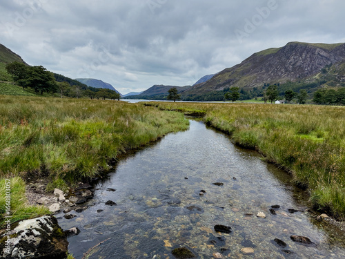 Photos A tranquil stream winding through grassy meadows leading to a mountain lake beneath a cloudy sky