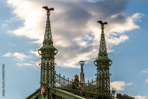 Photography Close-up of Freedom Bridge's upper spires with Turul birds against blue sky, showing Liberty Monument on Gellert Hill between them, highlighting Art Nouveau architecture and iconic golden spheres