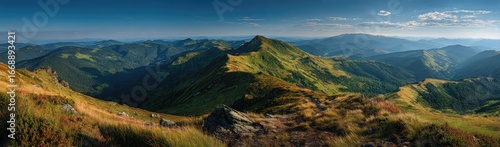 Panoramic vista of sunlit mountain range, grassy slopes, and distant hazy peaks under a clear blue sky