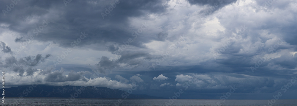 Obraz premium Stormy sky over mountains and sea. Dark dramatic clouds hang over the water surface, creating an atmosphere of tension and grandeur of nature. Podaca village, Makarska Riviera, South Dalmatia, Croatia
