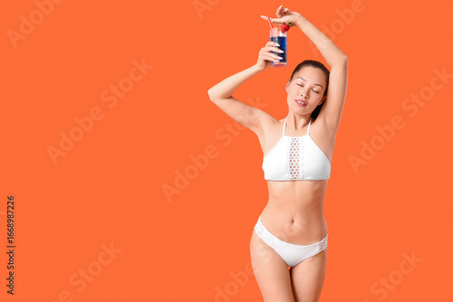 Beautiful young Asian woman in swimsuit with glass of cocktail on orange back...