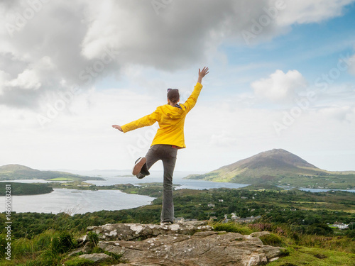 Canvastavla A woman in a yellow jacket is standing on a mountain top, looking out over a beautiful landscape