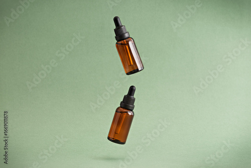Two amber dropper bottles with essential oil or cosmetic serum floating in the air on a green background. Concept of body care, rejuvenation. Close-up, copy space