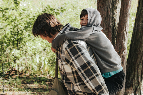 Wallpaper Mural A cheerful boy in a hoodie rides on the back of an older boy in a plaid shirt, sharing a playful piggyback moment outdoors in the sunlight. Torontodigital.ca