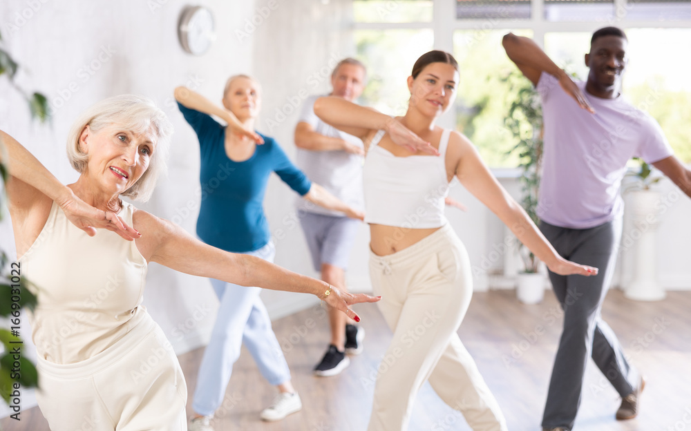 Obraz premium Young girl coach conducts classes for elderly in fitness studio. Female instructor teaches mature students of dance class basic movements of hip hop dance