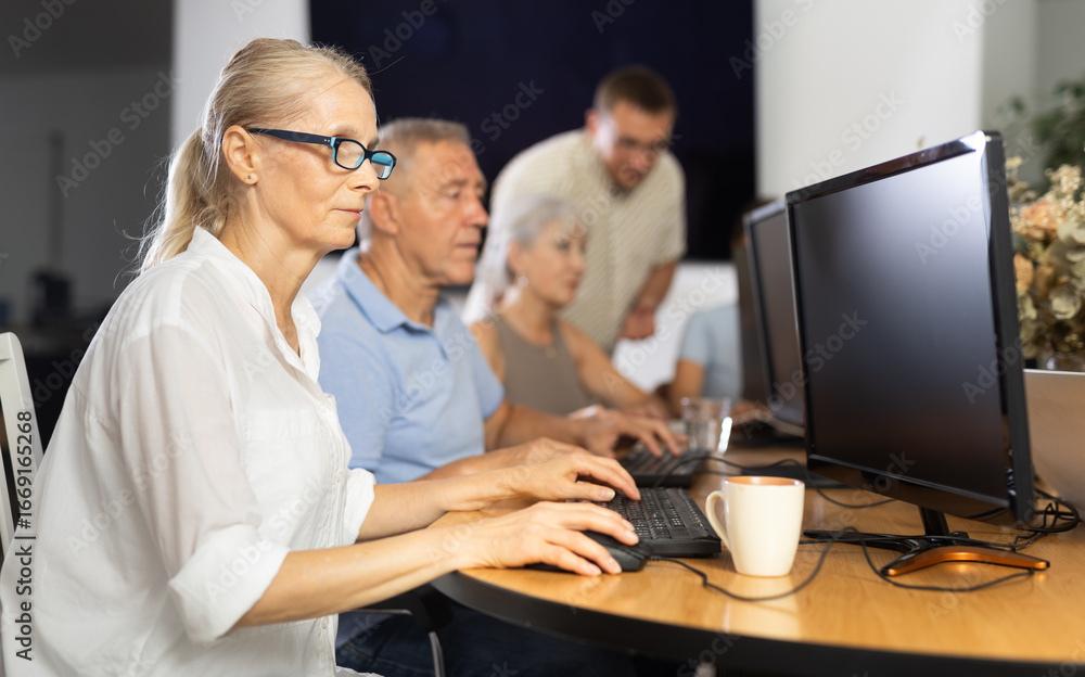 Obraz premium Motivated elderly woman in glasses mastering computer skills during group course with mentor standing on blurred background. Concept of curiosity of seniors towards technology