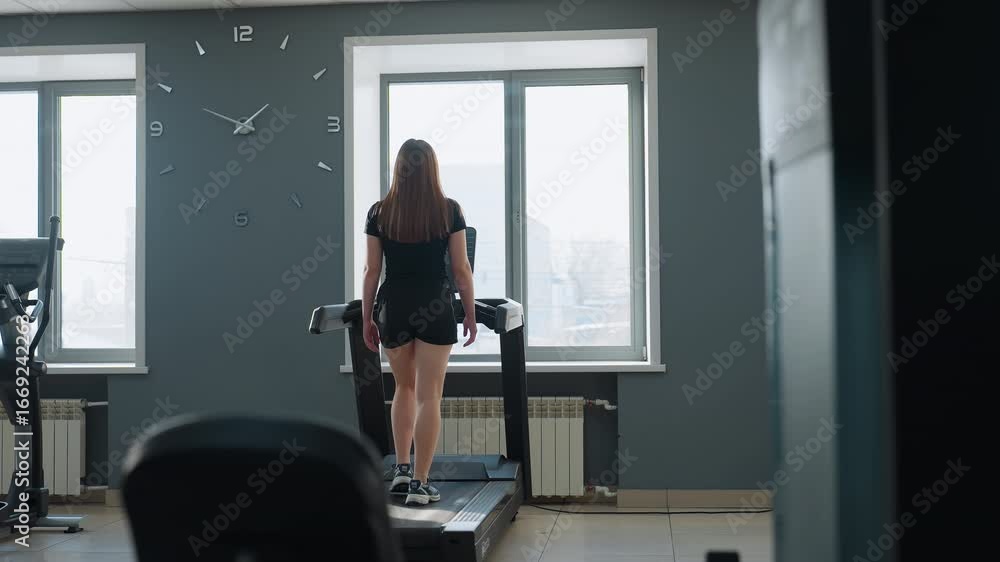 young woman in sportswear working out on treadmill facing window with sunlight streaming in, modern gym interior with second treadmill display blinking in background young woman in sportswear working out on treadmill facing window with sunlight streaming in, modern gym interior with second treadmill display blinking in background