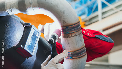 A technician in a bright red uniform works diligently on welding a metal pipe in an industrial setting. Bright lights illuminate the workspace, showcasing attention to detail and craftsmanship