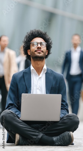 Person meditating, laptop on lap, outdoors. Other people walk by, blurred background