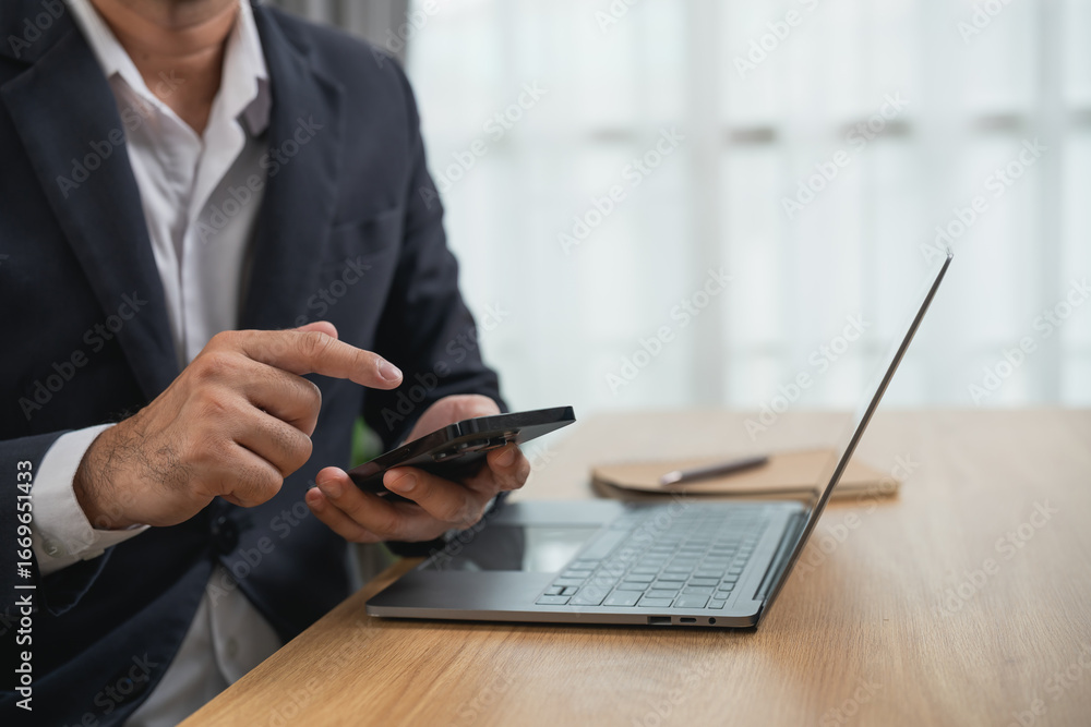 Fototapeta premium Business professional in suit using smartphone while sitting at wooden desk with laptop, notepad, and pen, focused on technology and productivity