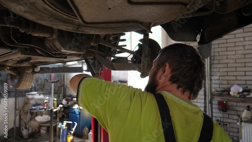 Male auto mechanic works underneath a lifting vehicle at workshop. Adult professional repairman fixing car at service or garage. Concept of regular automobile maintenance. Close up Slow motion