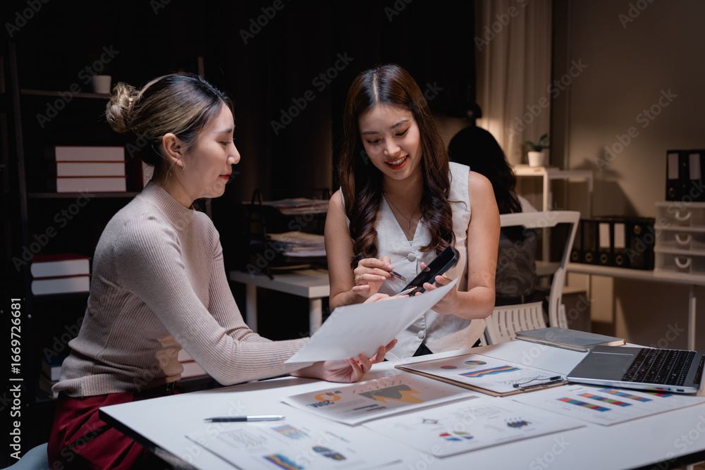 Fototapeta premium Two young Asian businesswomen are working late at night in the office, collaborating on a project, analyzing financial documents, using smartphone and laptop
