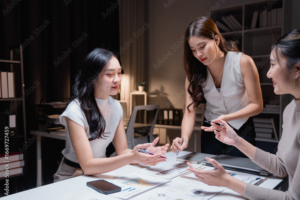 Fototapeta premium Three asian businesswomen are working late in the office, standing and sitting around a table, analyzing financial charts and discussing strategies during a productive meeting