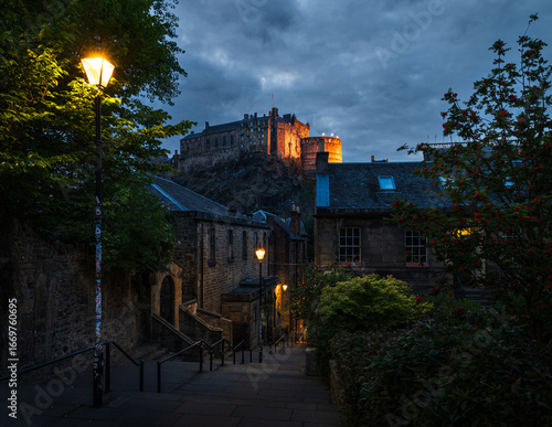 Blick vom Vennel Viewpoint in Edinburgh zur blauen Stunde auf Edinburgh Castle, gelb beleuchtete Burg auf dem Castle Rock, Fackeln auf den Zinnen und gelbe Straßenlaternen entlang der Gasse