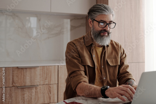 Horizontal medium portrait shot of handsome mature man with beard on face wearing eyeglasses sitting at table surfing Internet on laptop