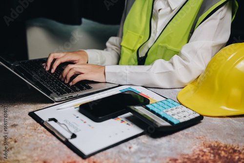Business professional in safety vest working on laptop with calculator, smartphone, and clipboard on desk
