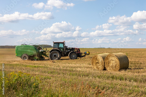 Round hay bales in field with baler wrapper machine working in background