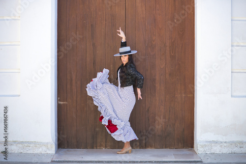Young woman, beautiful, brunette, flamenco dancer, wearing a beautiful dress with jacket and hat, dancing flamenco with a wooden door in the background. Flamenco concept, typical, Spanish, Seville.
